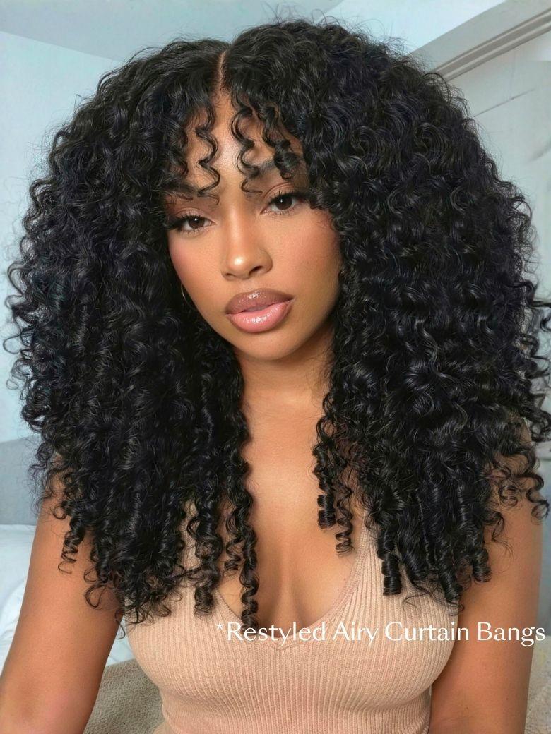 A woman with voluminous, defined curly hair and wispy bangs poses indoors, showcasing a gorgeous Burmese curly wig. She wears a beige top and natural makeup.