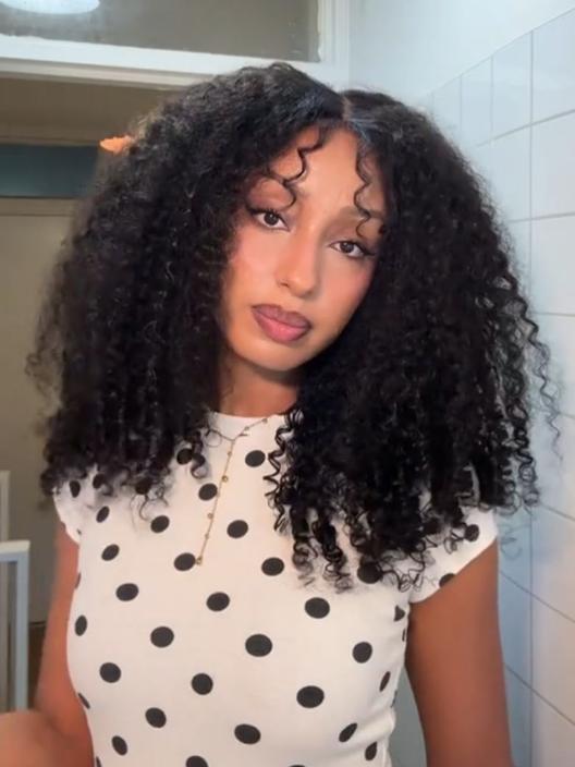 A woman with voluminous, defined curly hair and restyled wispy bangs poses indoors, showcasing a gorgeous Burmese curly wig. She wears a beige top and natural makeup.