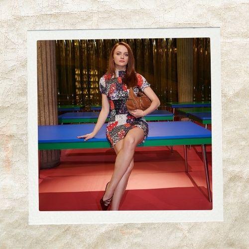 Joey King wearing a patterned mini dress with bright reddish-brown curly hair seated on a table