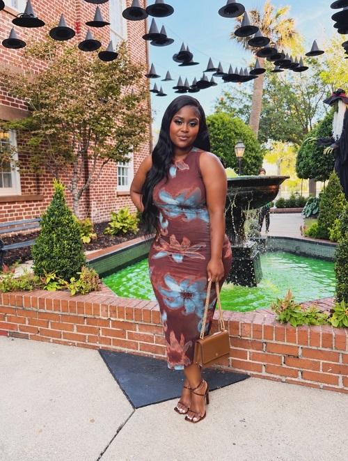 A woman in a patterned midi dress standing outdoors near a fountain, showcasing a long black lace front wig.
