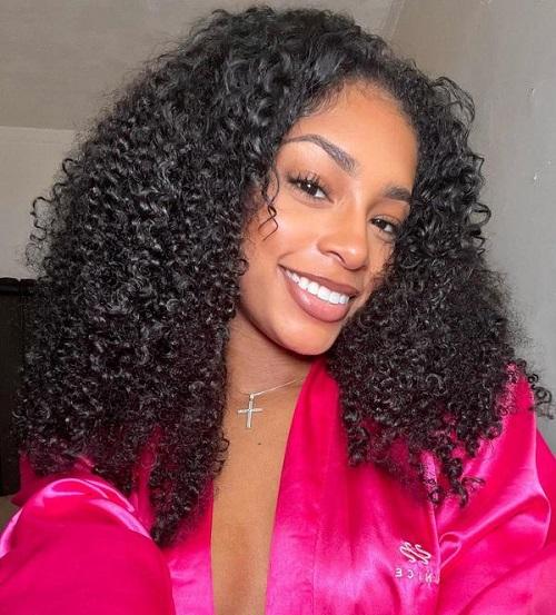A smiling woman wearing a pink silk robe, presenting long curly hair half wig.
